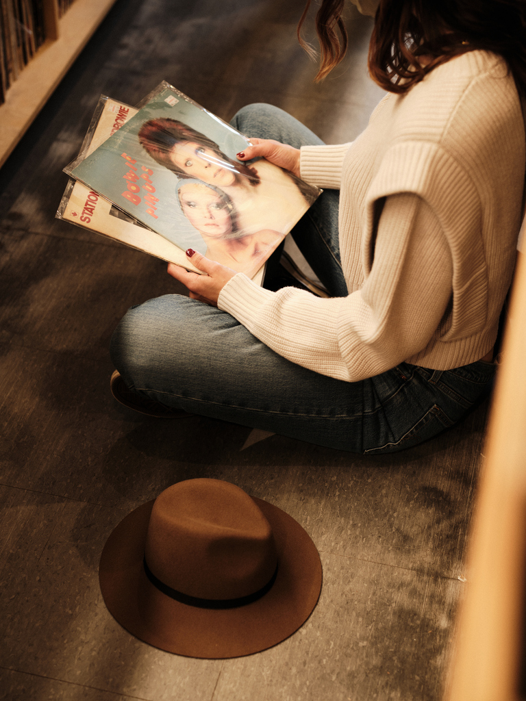 Person sitting on floor browsing vinyl records, wearing a cozy sweater, with a brown hat beside them in a record store.
