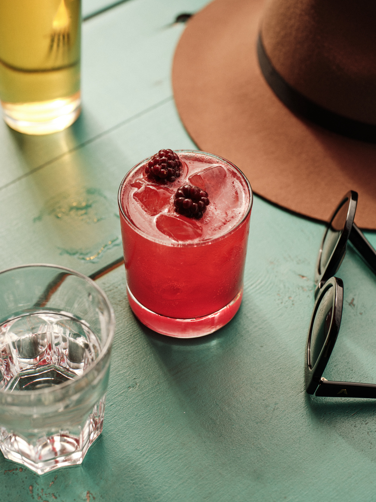 Refreshing red cocktail with blackberries on ice, beside a glass of water and sunglasses on a teal table.