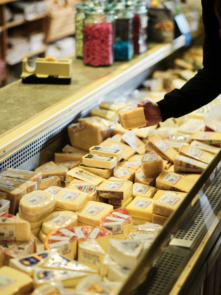 Person selecting cheese from a variety-filled deli counter, with jars of colorful candies in the background.