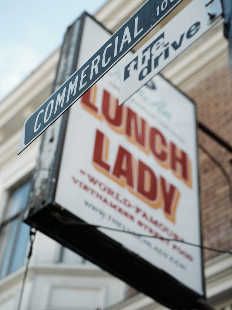 Street signs for Commercial Drive, with a blurred view of Lunch Lady restaurant sign in the background.