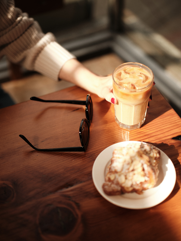 Iced coffee and pastry on a wooden table with sunglasses, hand reaching out in a cozy cafe setting.