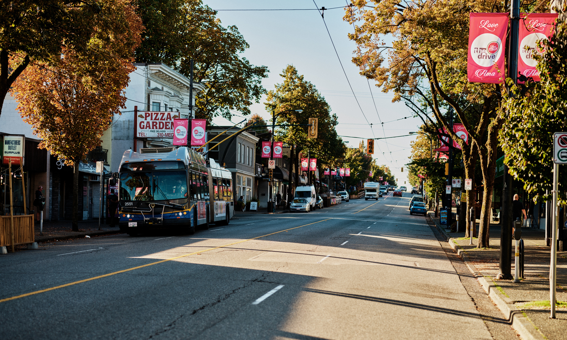 City street with public bus and autumn trees, vibrant banners lining the sidewalk, and shops in the background.
