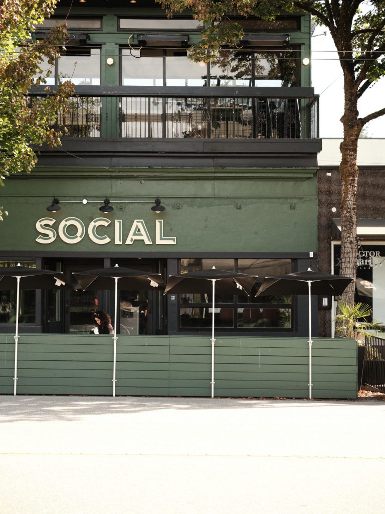 Green building with Social sign, outdoor seating under umbrellas, and trees lining the street.